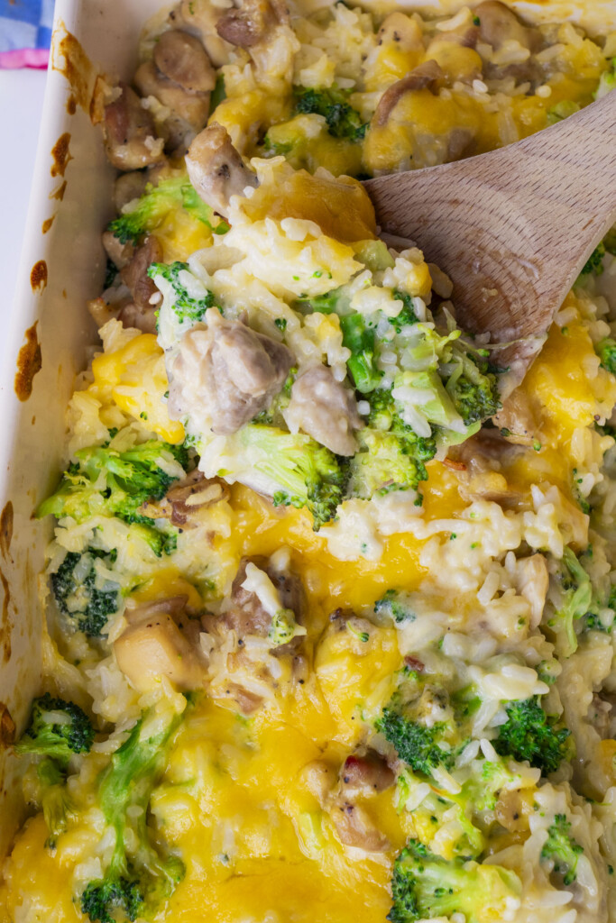 A close-up of a cheesy broccoli, chicken, and rice casserole being served with a wooden spoon from a baking dish. The dish is topped with melted VELVEETA cheese.