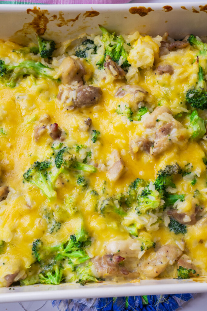 A close-up of a baked casserole in a white dish, featuring melted cheddar cheese, pieces of chicken, broccoli florets, and rice.
