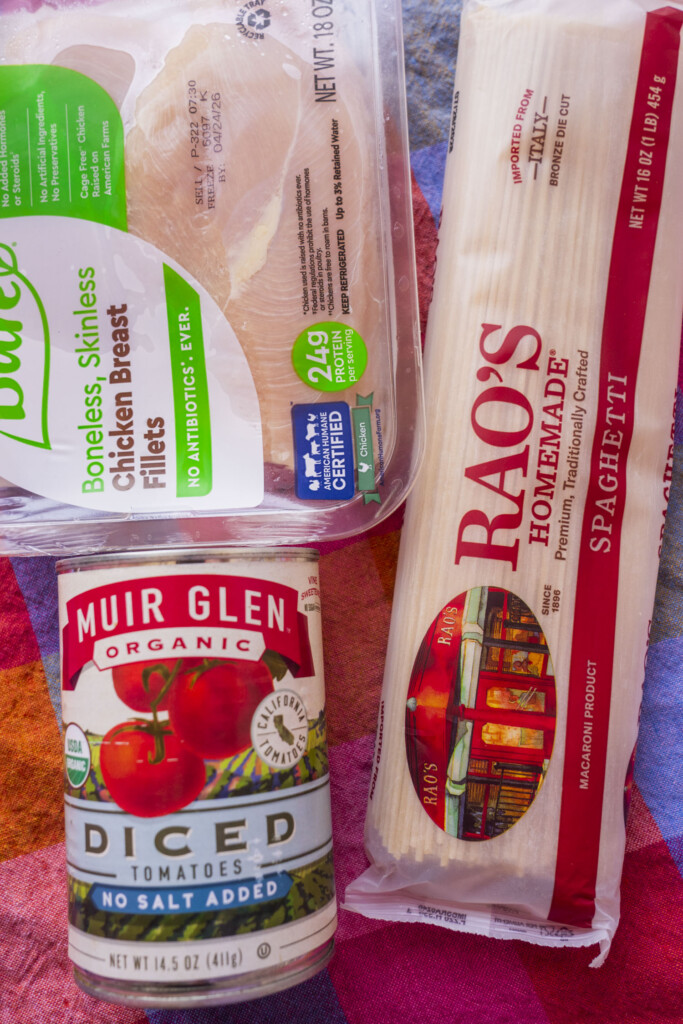 A package of boneless, skinless chicken breasts, a box of Rao’s spaghetti, and a can of Muir Glen organic diced tomatoes are arranged together on a pink and purple tablecloth.