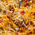 Close-up of baked spaghetti mixed with melted cheddar cheese, chunks of chicken, and pieces of tomato, with a serving utensil visible among the noodles.