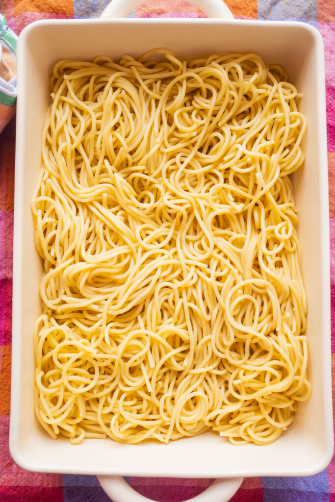 A white rectangular baking dish filled with cooked plain spaghetti noodles, placed on a colorful, checkered cloth.