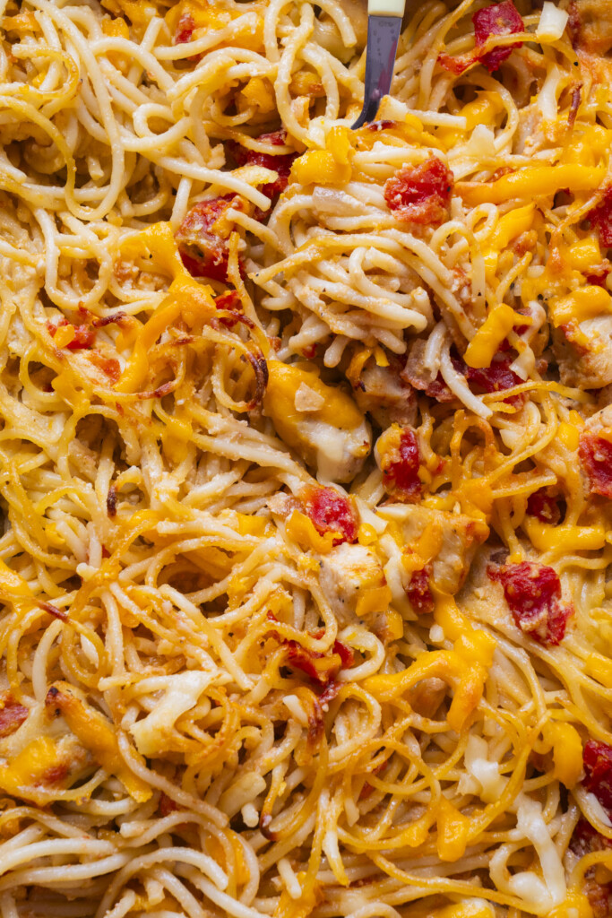 Close-up of baked spaghetti mixed with melted cheddar cheese and diced tomatoes, with a fork lifting a portion from the dish.