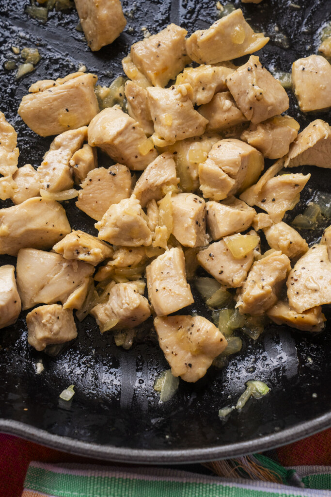 Cooked, diced chicken pieces seasoned with black pepper and sautéed onions in a black skillet. The chicken is golden brown and glistening, with some visible oil and browned bits on the pan.