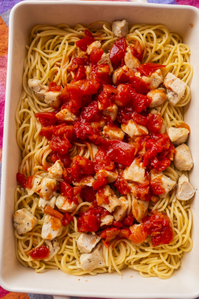 A rectangular dish filled with cooked spaghetti noodles, topped with chunks of cooked chicken and diced tomatoes in a red sauce.