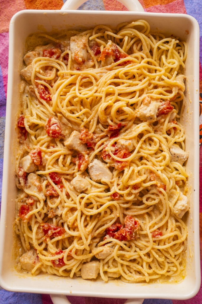 A rectangular white dish filled with spaghetti mixed with chunks of chicken and diced tomatoes in a creamy sauce sits on a colorful tablecloth.
