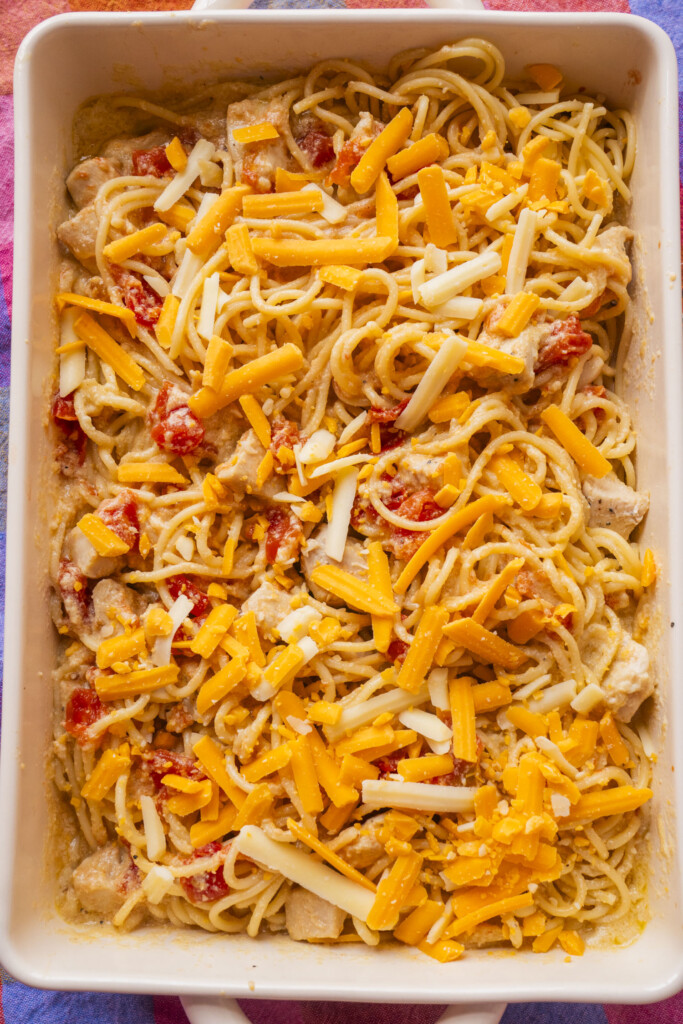Baked spaghetti in a rectangular dish, topped with shredded cheddar and mozzarella cheese, visible pieces of chicken, and diced tomatoes mixed throughout.