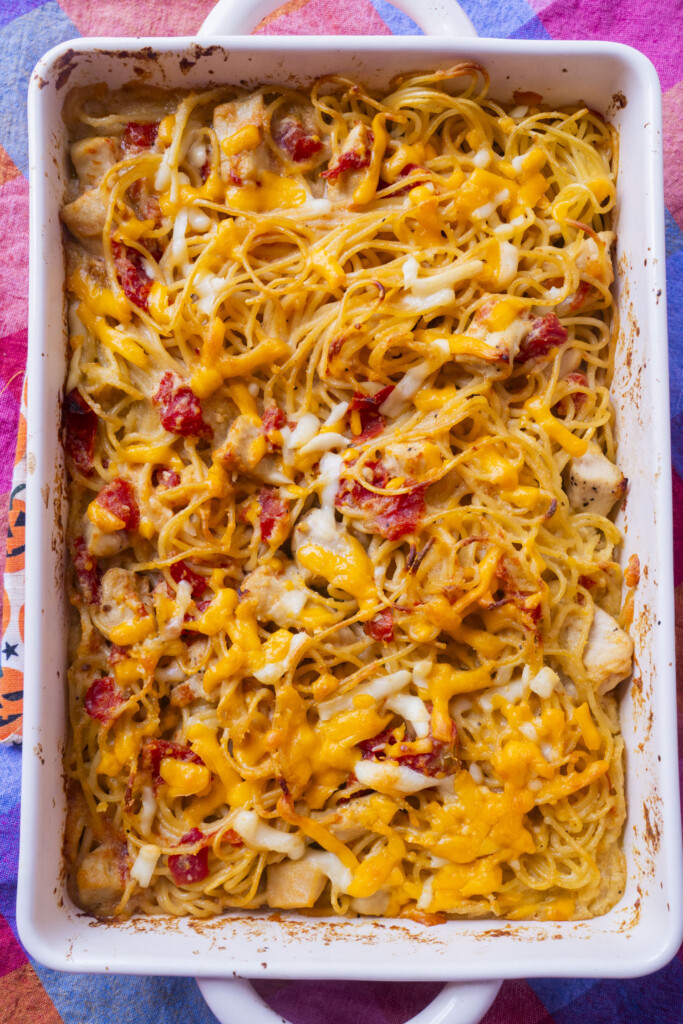 A white baking dish filled with baked spaghetti casserole topped with melted cheddar cheese, pieces of chicken, and bits of tomato, set on a colorful tablecloth.