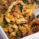 A close-up of a baked pasta casserole with rotini noodles, ground meat, spinach, tomato sauce, and melted cheese being scooped with a wooden spoon.