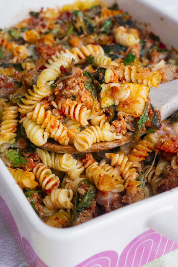 A close-up of a baked pasta casserole with rotini noodles, ground meat, spinach, tomato sauce, and melted cheese, served in a white dish with a wooden spoon scooping a portion.