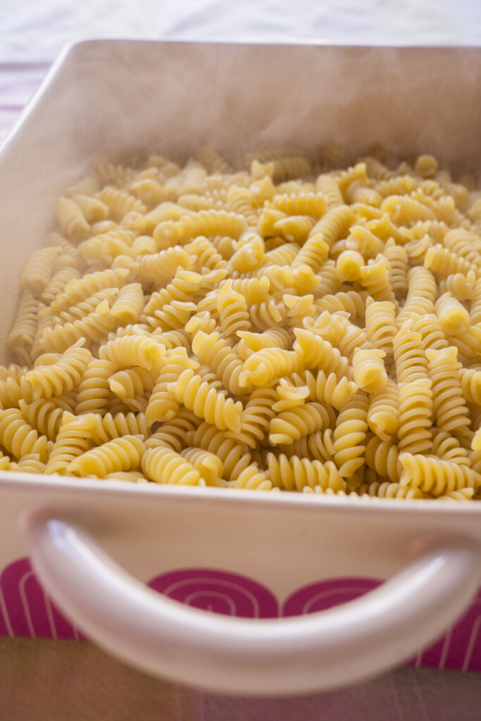 A close-up of freshly cooked spiral pasta (rotini) in a white ceramic baking dish, with steam rising from the noodles, indicating that they are hot and ready to be served.