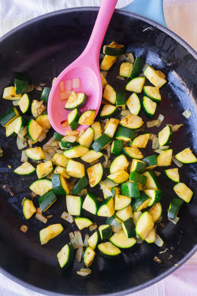 Chopped zucchini and onions are sautéing in a black skillet, stirred with a pink spoon. The vegetables appear lightly browned and cooked.