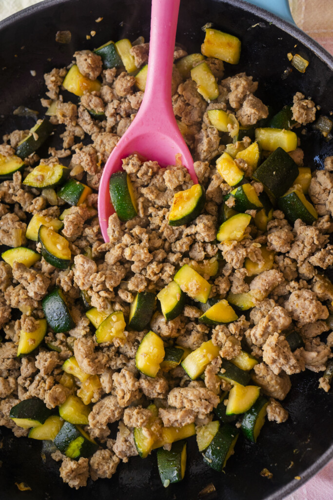 A skillet filled with cooked ground meat and chopped zucchini pieces, being stirred with a pink spoon.
