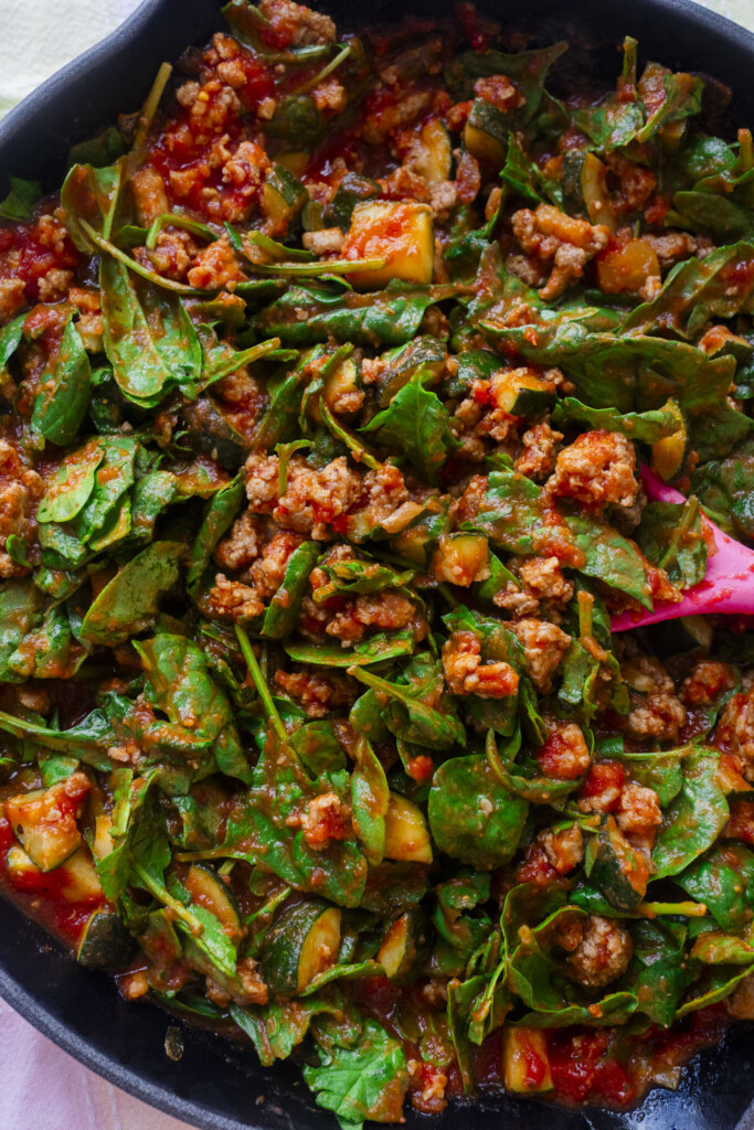 A close-up of a skillet filled with cooked ground meat, fresh spinach leaves, tomato sauce, and diced vegetables being stirred with a pink spatula.