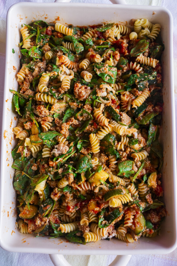 A rectangular white baking dish filled with rotini pasta, ground meat, spinach, tomato sauce, and zucchini, mixed together for a colorful pasta bake.