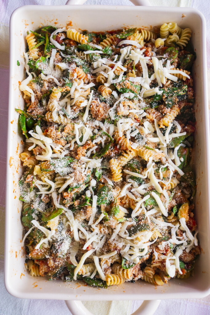 A rectangular baking dish filled with rotini pasta, tomato sauce, spinach, and ground meat, topped with shredded mozzarella and grated Parmesan cheese.