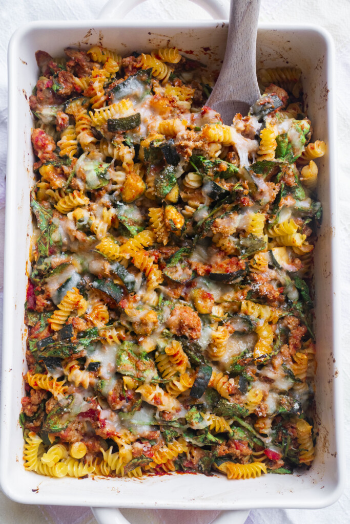 A baked pasta casserole with rotini, zucchini, spinach, tomato sauce, melted cheese in a white dish, with a wooden spoon in the corner.