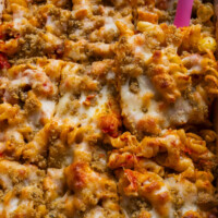 Close-up of baked pasta topped with melted cheese and golden brown breadcrumbs, with a pink spatula lifting a portion from the dish.
