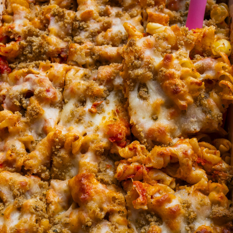 Close-up of baked pasta topped with melted cheese and golden brown breadcrumbs, with a pink spatula lifting a portion from the dish.