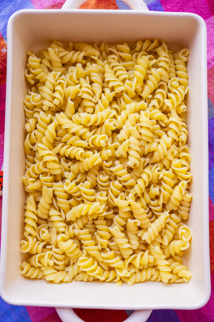 A white rectangular baking dish filled with cooked rotini pasta, placed on a colorful, patterned tablecloth.