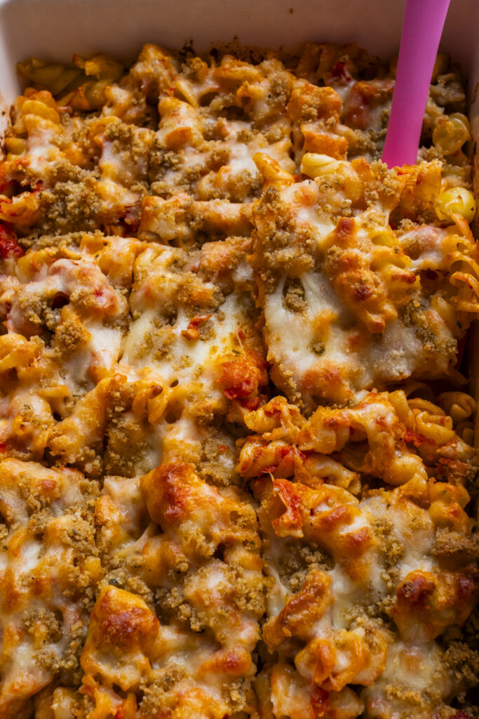 Close-up of a cheesy baked pasta casserole with melted cheese and golden breadcrumbs on top. The dish is cut into squares, and a pink spatula is visible for serving.