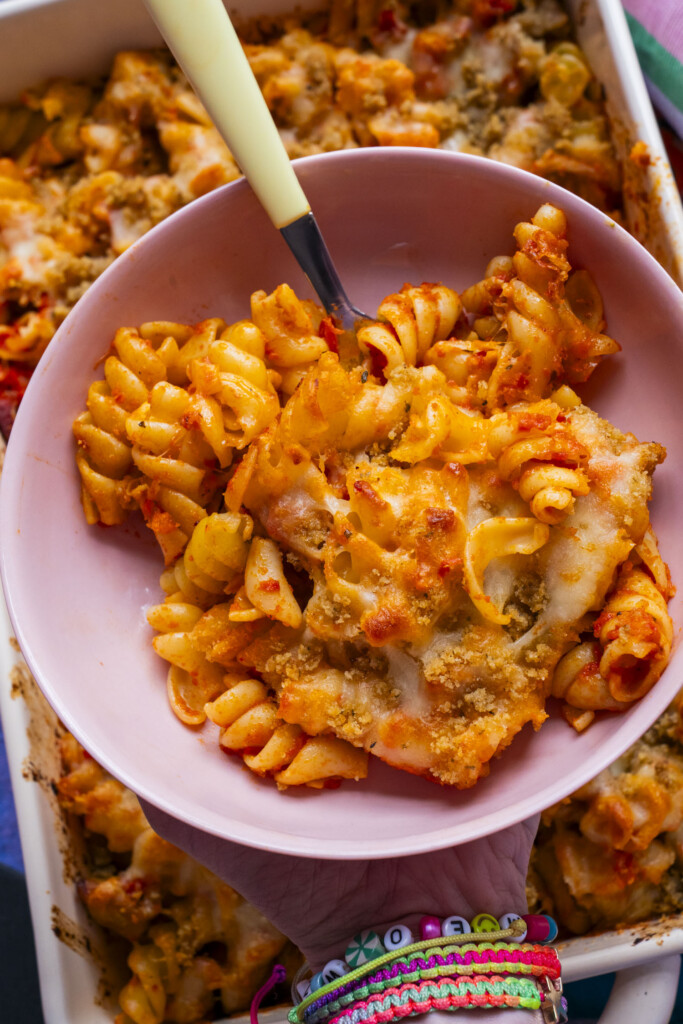 A hand holding a pink bowl filled with cheesy baked rotini pasta topped with breadcrumbs, with a baking dish of more pasta in the background. The person wears colorful beaded bracelets.