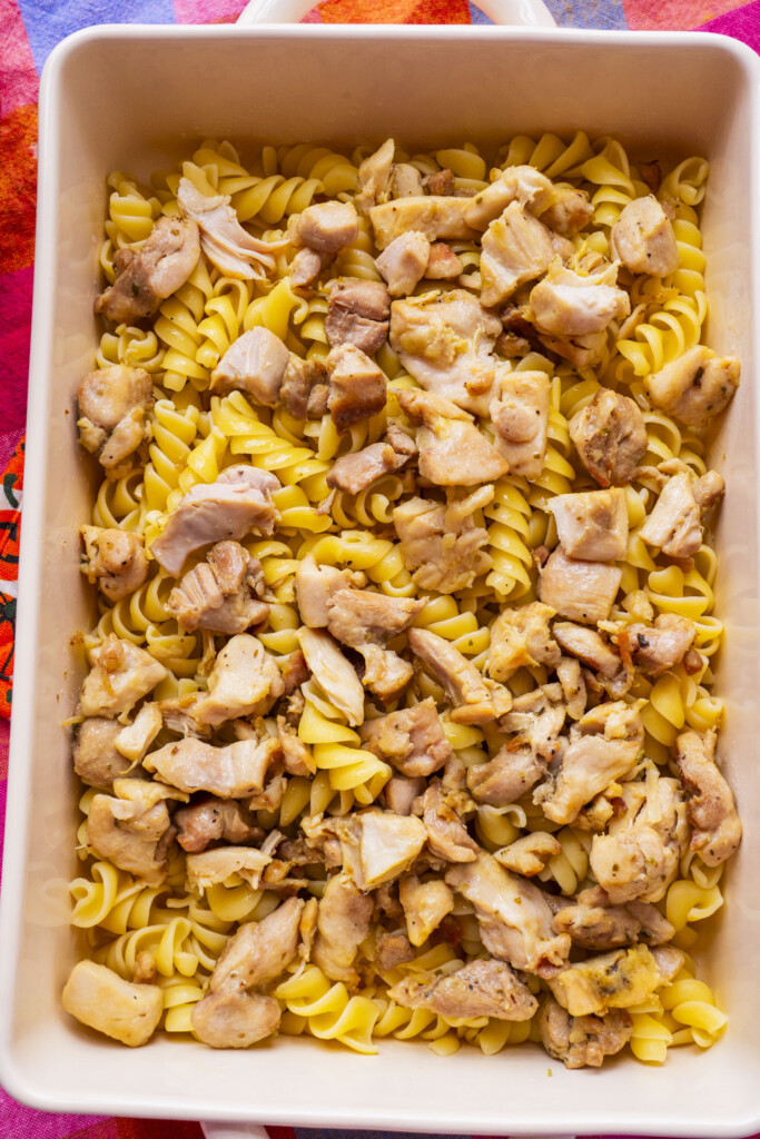 A baking dish filled with cooked rotini pasta topped with pieces of cooked chicken, seen from above on a colorful surface.
