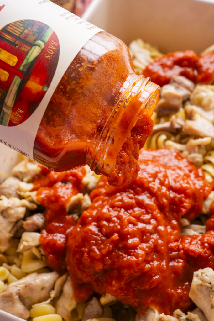 A close-up of Rao's pasta sauce being poured from a jar onto a dish filled with pasta and pieces of cooked chicken.