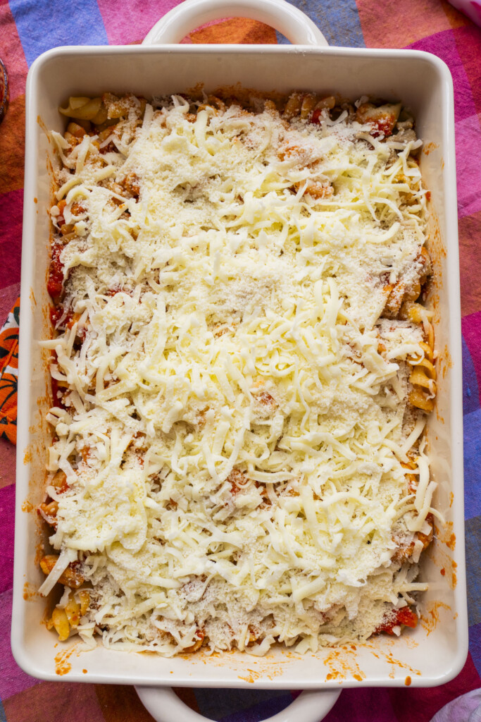 A rectangular white baking dish filled with an uncooked pasta bake, topped with shredded cheese and grated Parmesan, sitting on a colorful tablecloth.