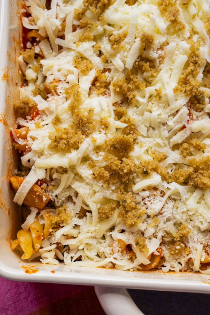 Close-up of an unbaked pasta casserole in a white dish, topped with tomato sauce, shredded cheese, grated parmesan, and golden brown bread crumbs.