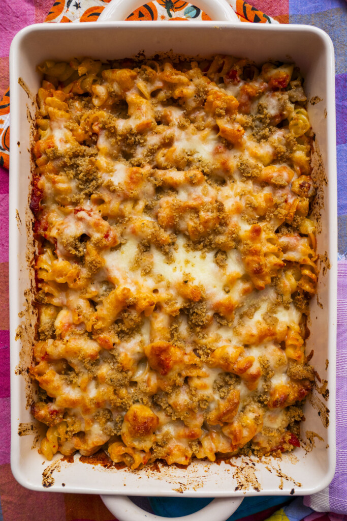 A baked pasta casserole in a rectangular white dish, topped with melted cheese and golden breadcrumbs, with visible browned edges around the sides. The dish sits on a colorful, patterned tablecloth.