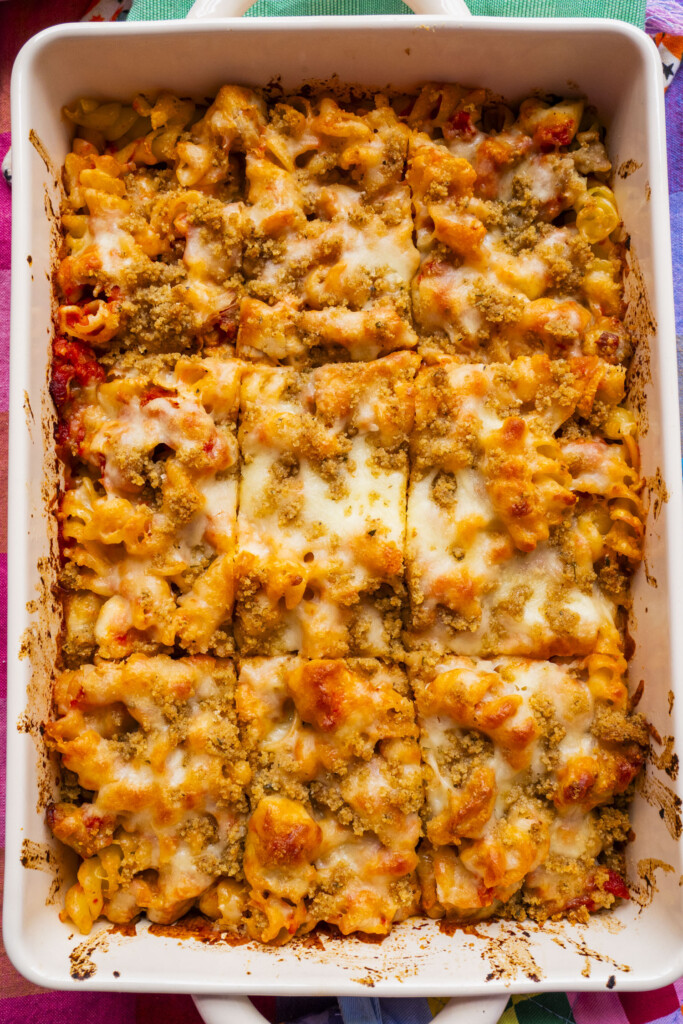 A baked pasta casserole in a white dish, cut into 9 portions. The top is golden brown with melted cheese and breadcrumbs, and the curly noodles and tomato sauce are visible.