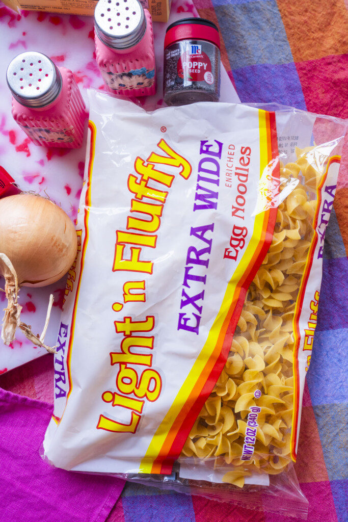 A bag of Light n Fluffy Extra Wide Egg Noodles sits on a colorful tablecloth, surrounded by a yellow onion, a jar of poppy seeds, and two pink salt and pepper shakers.