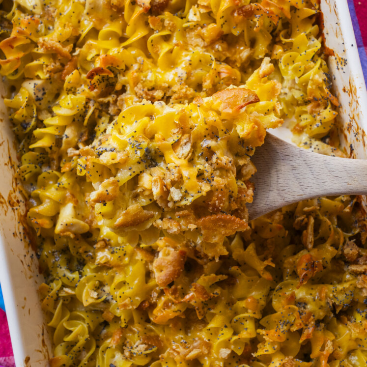 A close-up of a baked casserole dish filled with cheesy, golden-brown spiral pasta, topped with crispy breadcrumbs and poppy seeds. A wooden spoon is lifting a serving from the dish.