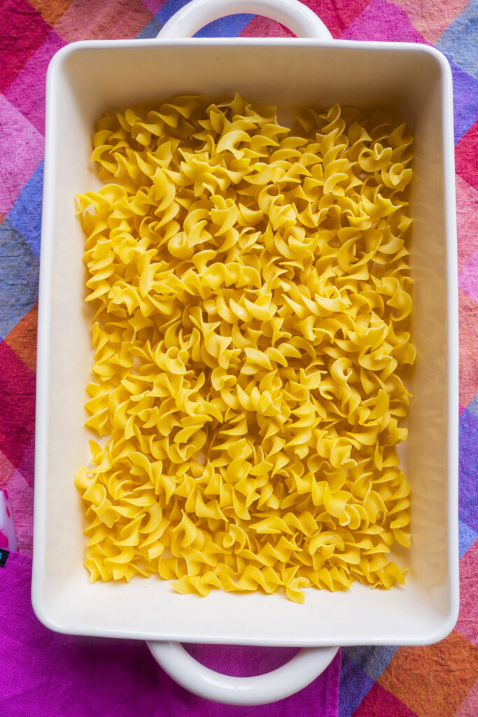 A white rectangular baking dish filled with uncooked egg noodles sits on a colorful, checkered tablecloth in shades of pink, orange, and blue.