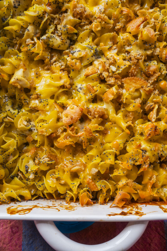 Close-up of a baked casserole dish filled with cheesy egg noodles, orange cracker crumbs, poppy seeds, and pieces of chicken, all topped with a browned layer of melted cheese.