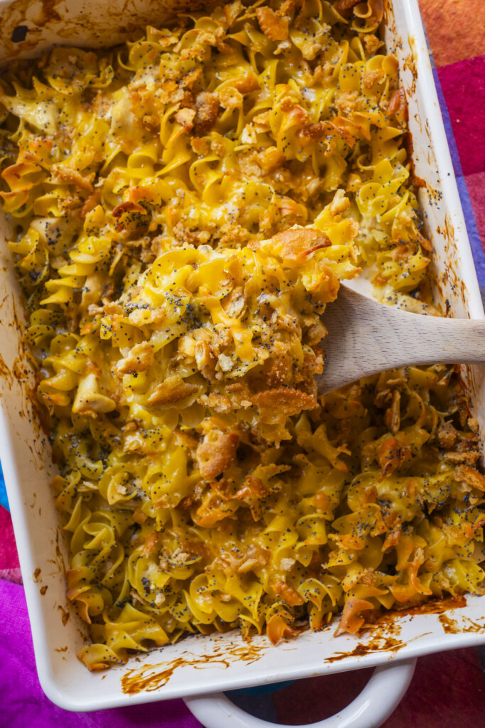 A wooden spoon scoops a cheesy baked noodle casserole topped with crispy crumbs and sprinkled with poppy seeds from a white baking dish.