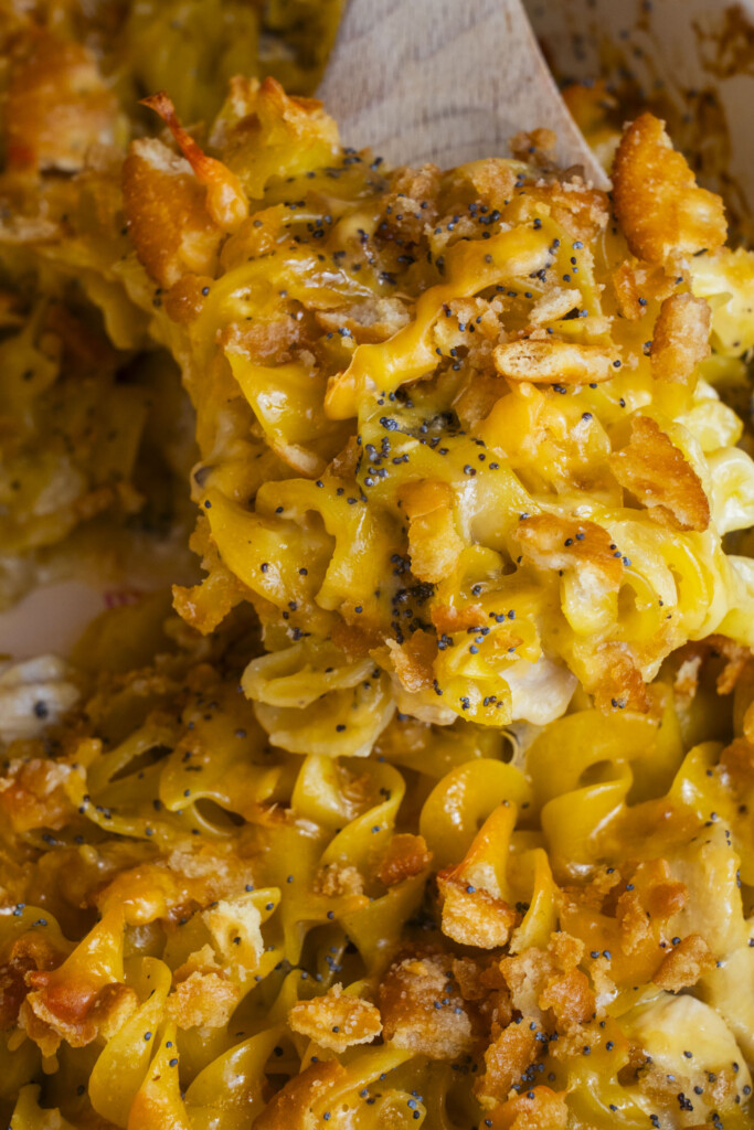 A close-up of a serving of cheesy baked noodle casserole with golden melted cheese, crispy breadcrumbs, and sprinkled black poppy seeds on top, being lifted with a wooden spatula.