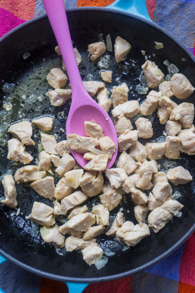 Chunks of chicken being cooked in a black skillet with a pink spoon, surrounded by colorful fabric.