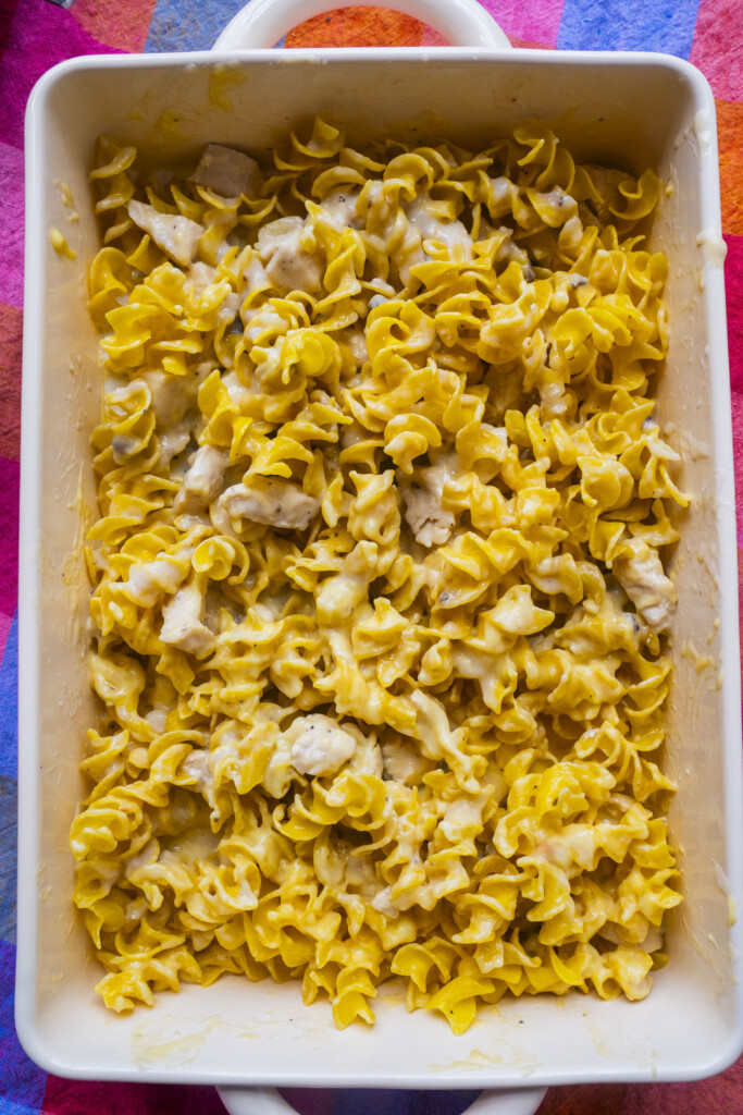 A white baking dish filled with cheesy pasta and chunks of chicken, with curly egg noodles coated in a creamy yellow sauce, sitting on a colorful, striped surface.
