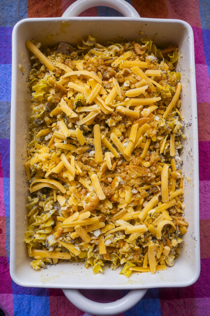 A rectangular baking dish filled with an unbaked casserole topped with shredded cheddar cheese and crushed crackers, sitting on a colorful checkered tablecloth.