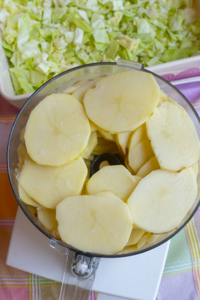 A food processor bowl filled with thinly sliced potatoes sits on a table, with chopped cabbage visible in a dish in the background.