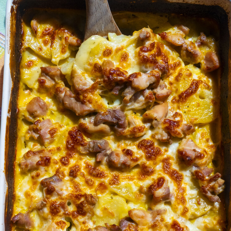 A close-up of a baked casserole with golden-brown cheese, pieces of chicken, and sliced potatoes. A wooden spoon is lifting a serving from the dish.