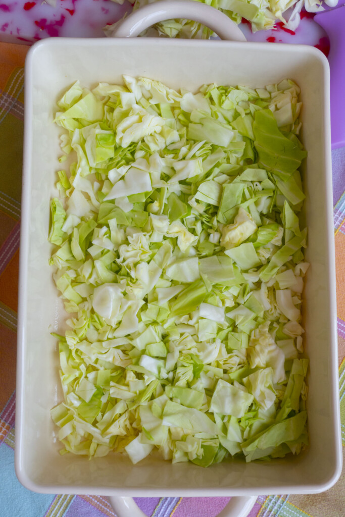 Chopped green cabbage spread evenly in a rectangular white baking dish, placed on a pastel-colored plaid tablecloth.