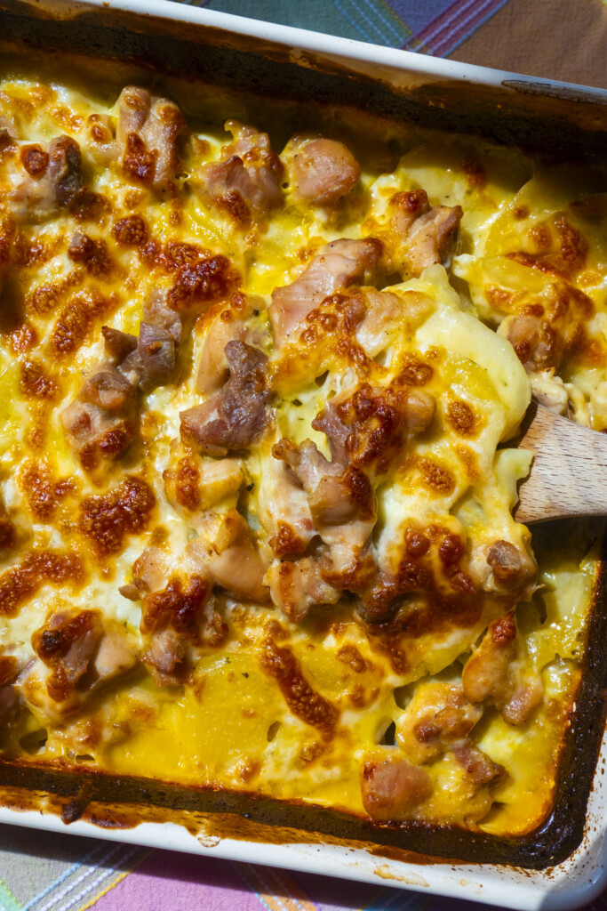 A close-up of a baked casserole with golden, melted cheese on top, visible pieces of browned chicken, and creamy sauce. A wooden spatula is lifting a serving from the dish.