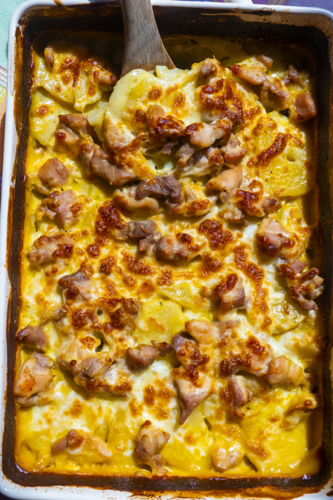Creamy, cheesy baked casserole with chunks of browned chicken  thighs and potatoes, being scooped with a wooden spoon. The dish has a golden, bubbly surface.