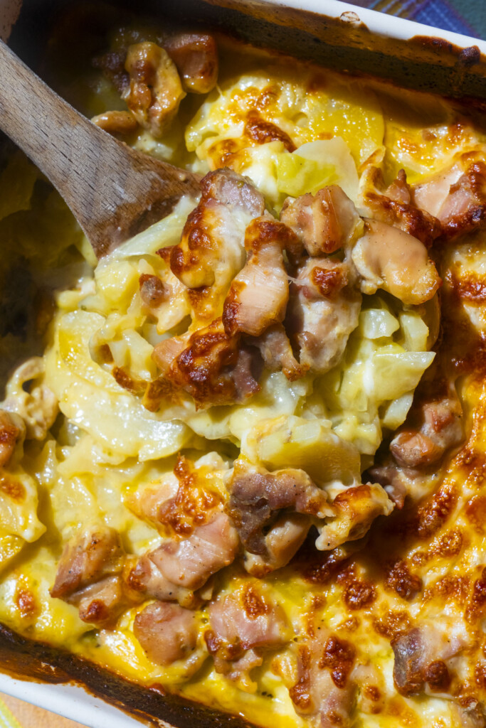 A close-up of a creamy, cheesy baked casserole with chunks of browned chicken and potatoes, being scooped with a wooden spoon. The dish has a golden, bubbly surface.