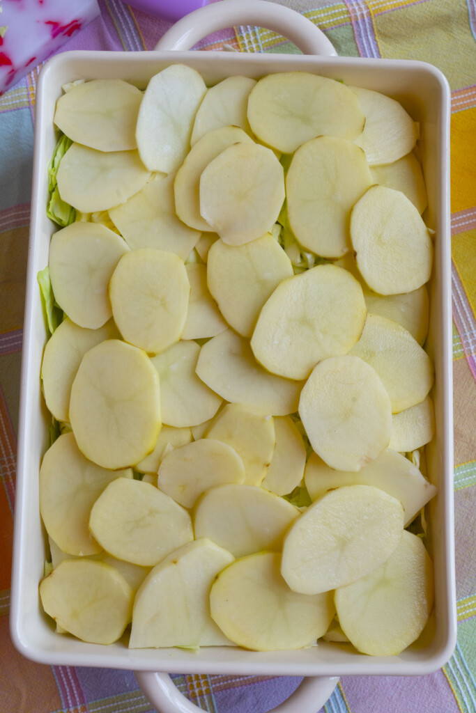 A rectangular baking dish filled with thinly sliced raw potatoes arranged in overlapping layers, set on a pastel-colored checkered tablecloth.