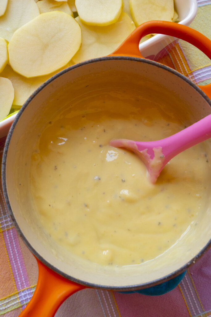 A pink spatula stirs creamy cheese sauce in an orange pot. In the background, a dish with thinly sliced potatoes sits on a colorful plaid tablecloth.