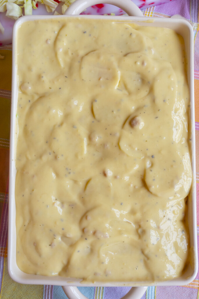 A rectangular baking dish filled with sliced potatoes covered in a creamy, yellow cheese sauce, ready to be baked. The dish sits on a pastel-colored plaid cloth.