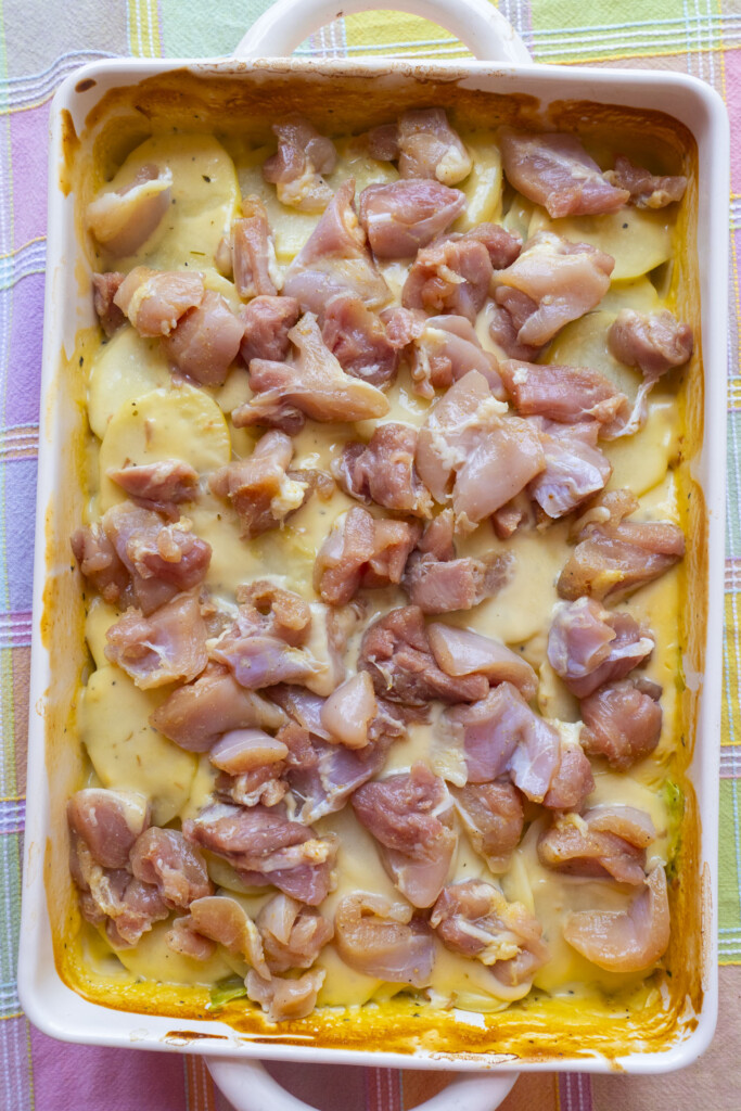 Casserole dish filled with sliced potatoes, creamy sauce, and uncooked chunks of chicken, ready to be baked. The dish sits on a pastel plaid tablecloth.